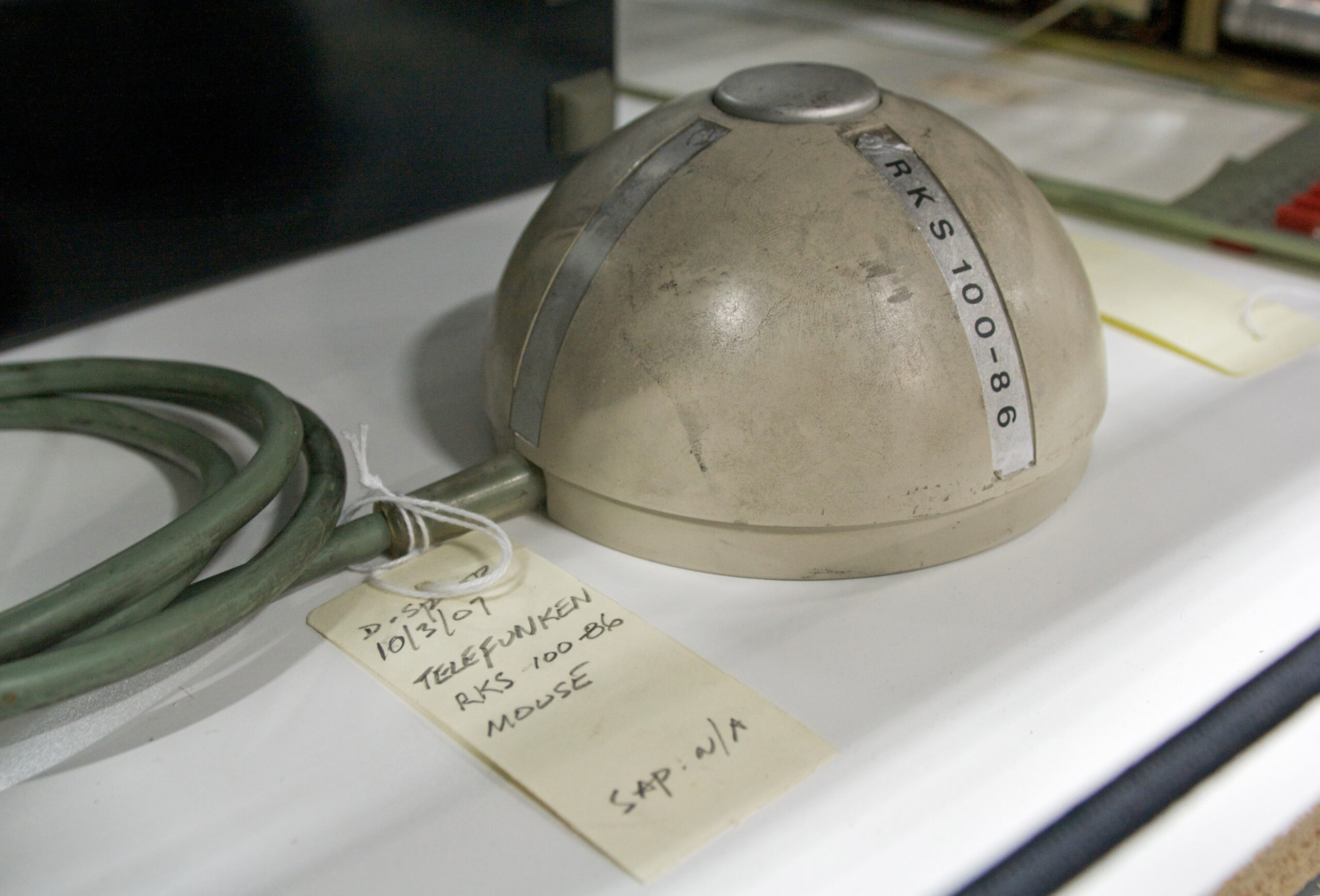 Photograph of an early rolling-ball mouse from the 1960s, shaped like half a cylinder and sitting on a desk with a label.