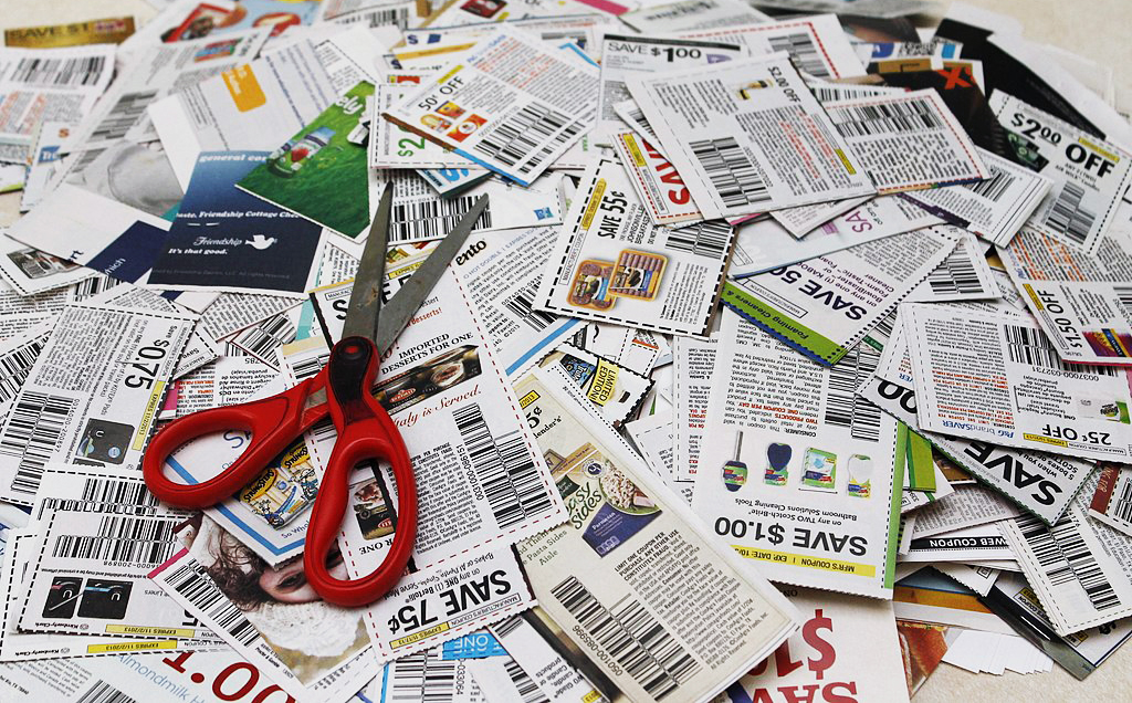 Photograph of a pair of red scissors sitting on top of a large pile of coupons.