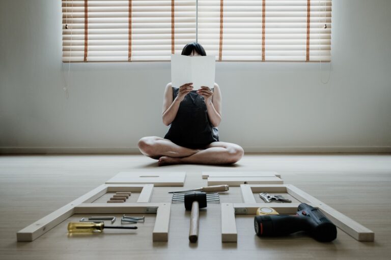 Photograph of a person sitting cross-legged, reading something very close to their face, with many tools and pieces of wood laid out in front of them.