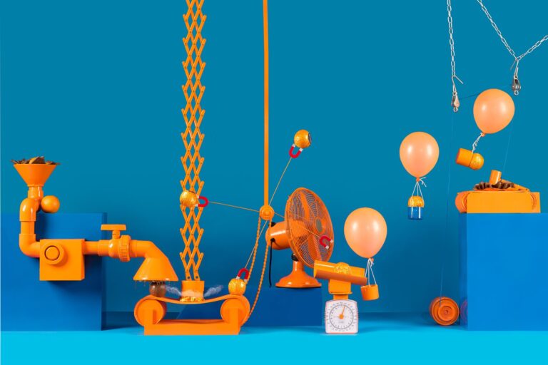 A colorful machine made up of different orange contraptions including an oscillating fan and balloons, with a rich blue background.