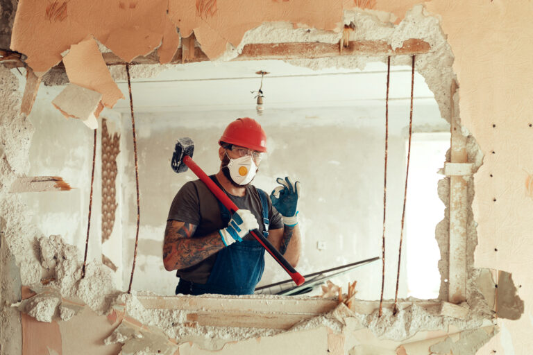 Image of a construction worker with a sledgehammer giving the "OK" sign through a smashed hole in a wall.