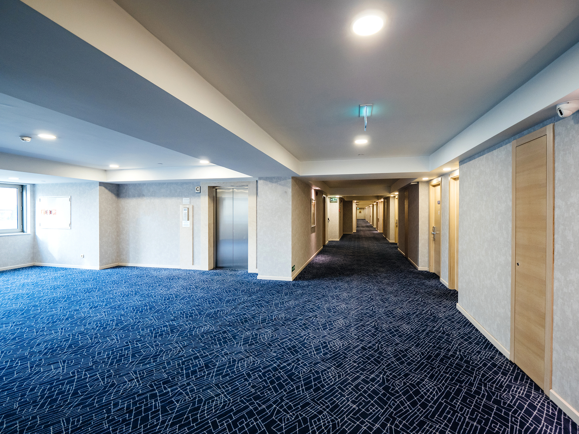 Photograph of a hallway and elevator bank of a floor in a hotel, with dark blue carpet and off-white walls.