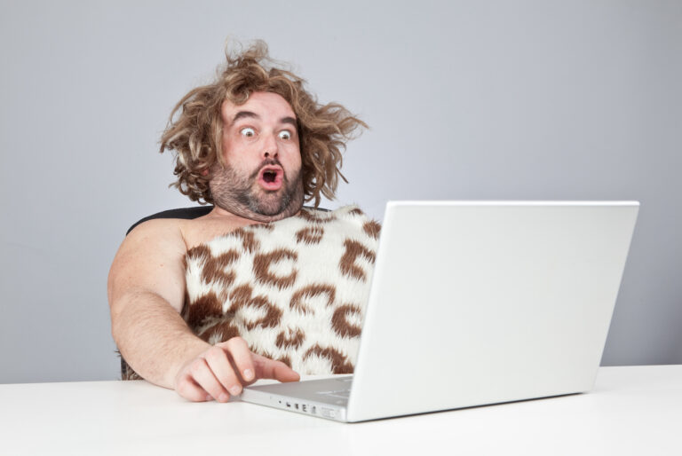 Stock image of a "caveman" dressed in an animal-skin garment looking shocked at a computer.