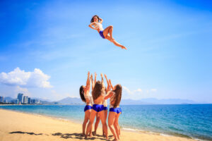 Photograph of a group of cheerleaders on a beach tossing one cheerleader high in the air.