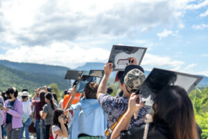 A group of people standing in a mountainous area using reflective surfaces to look at an eclipse.