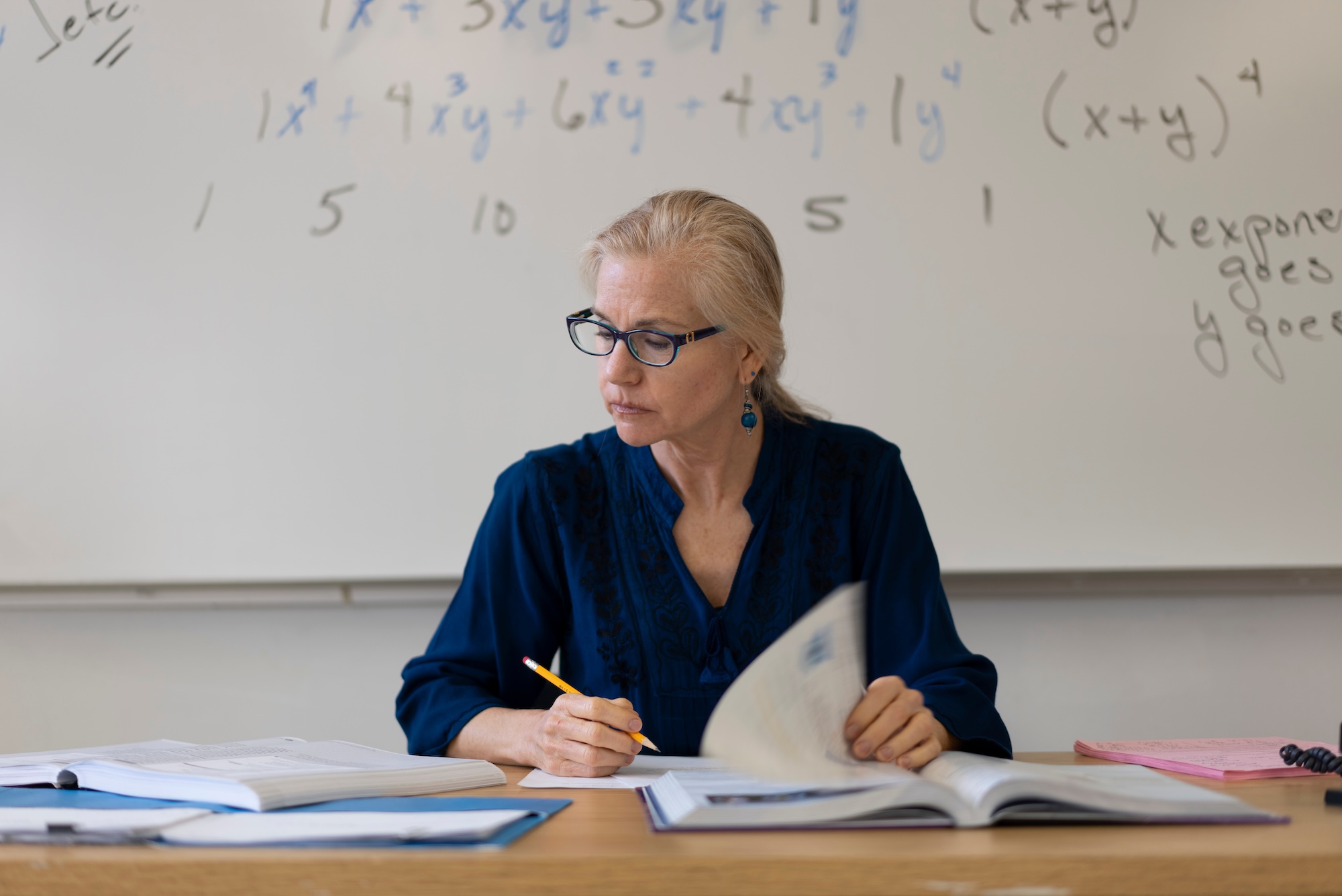 Photograph of a teacher sitting at a desk grading papers in front of a board with math formulas.