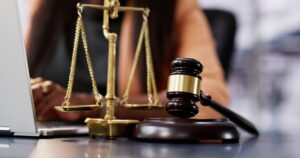 Photograph a "lawyer"'s desk featuring a small gavel and scales of justice, with a figure on a computer in the background.