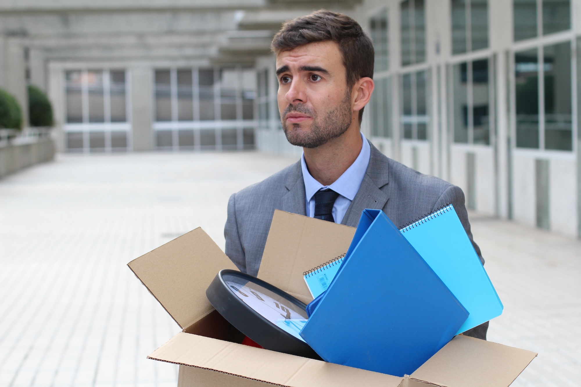 Photograph of a man who has just been fired, walking in front of an office building, a cardboard box with his things (including two blue folders and an analog clock) in his arms.