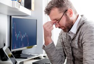 Stock image of a man pinching his nose in sadness in front of a screen showing a sharply declining line graph.