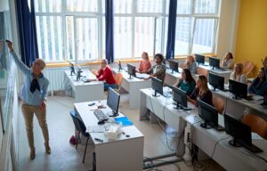 Photograph of a classroom with students seated in front of computers and a teacher at the board.