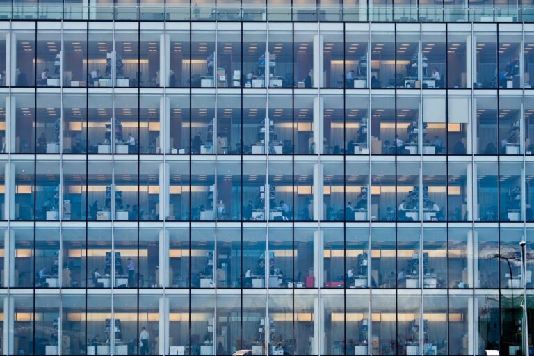 Photograph from the exterior of a large office building with fully glass sides showing many floors of cubicles.