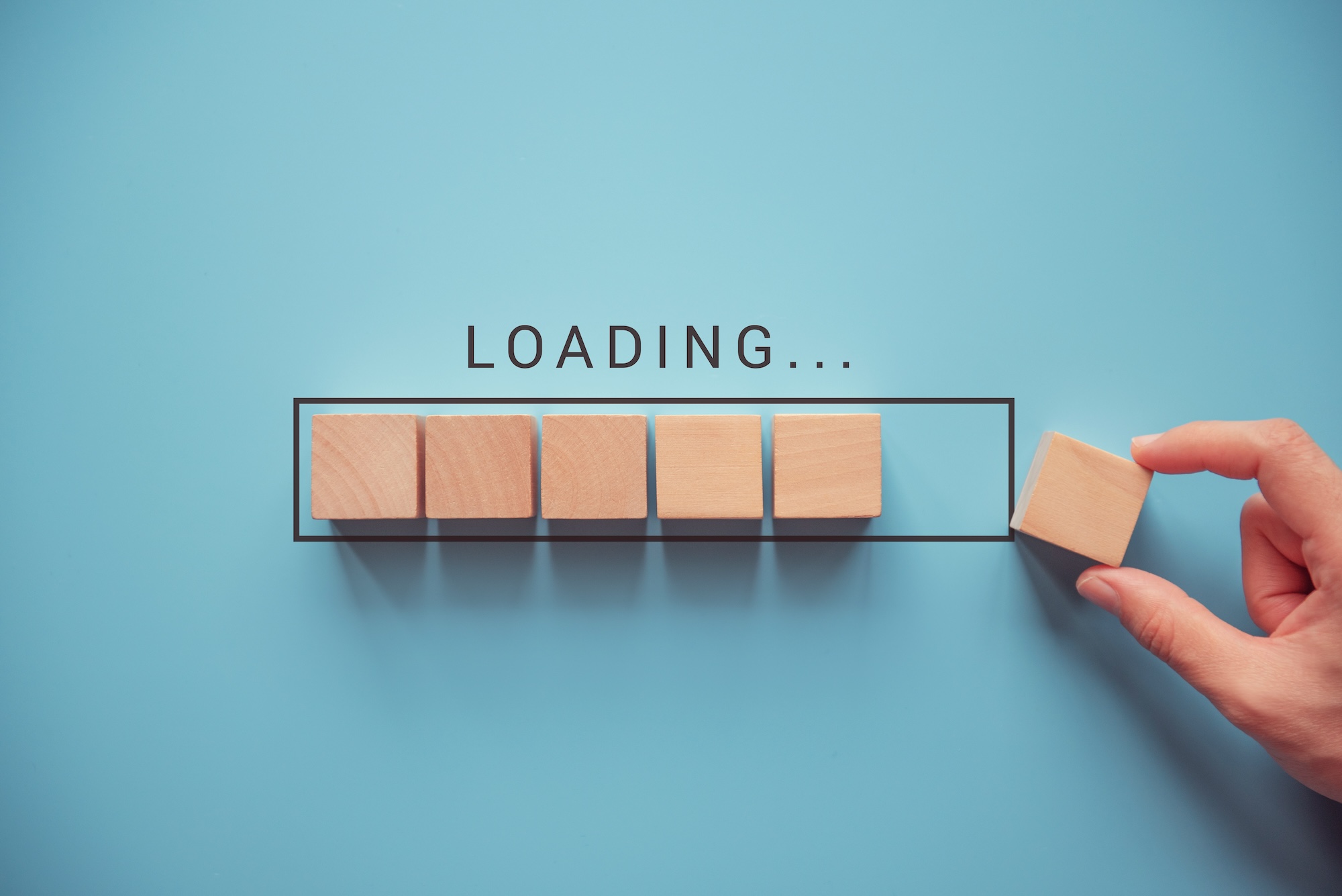 Image of several wooden blocks imitating a loading bar with a hand placing the final block. The word "LOADING" above it. Blue background.