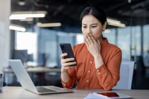 Image of a woman sitting at a desk in front of a laptop, holding a mobile phone and pressing her other hand against her mouth in concern.