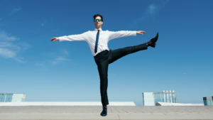 Photograph of a man in a business suit (without jacket) and sunglasses on the roof of a building, doing a dance pose with his arms straight out and one leg outstretched above 90 degrees.