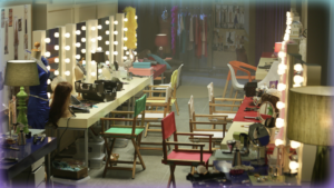 Image of a backstage area with many mirrors lined with big bulb lights and chairs in front of them.