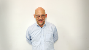 Image of Clay wearing glasses and a button-down shirt, standing against a white background with his hands behind his back.