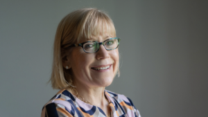 Headshot of Perry: a fair-skinned woman with blond hair (and bangs), wearing glasses and a multicolored shirt, standing against a grey background.