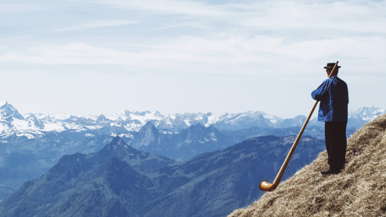 Image of a person standing on an Alpine peak blowing a long horn.