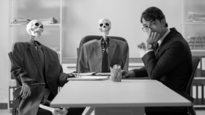 Black and white image of a man and two skeletons, all in suits, sitting at a board room table.