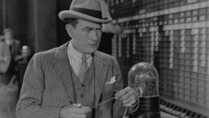 Black and white film still from The Silent Partner featuring a man in a suit and hat holding a paper from a 1920s-era stock ticker.