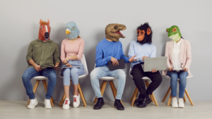 Image of five individuals dressed in casual clothing with animal-head masks on: from left to right, a horse, a pigeon, a dinosaur, a gorilla, and a lizard. Everyone holds a laptop or tablet and the gorilla's is open.