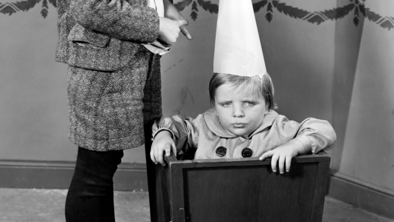 Black and white image of an unhappy looking child wearing a dunce cap.