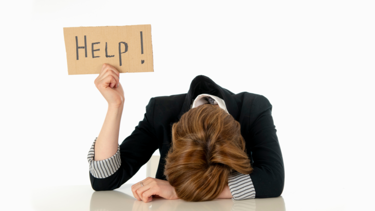 Image of a person in a blazer with their head on the arm that's resting on the table (in despair), holding up a sign that says, "Help!"