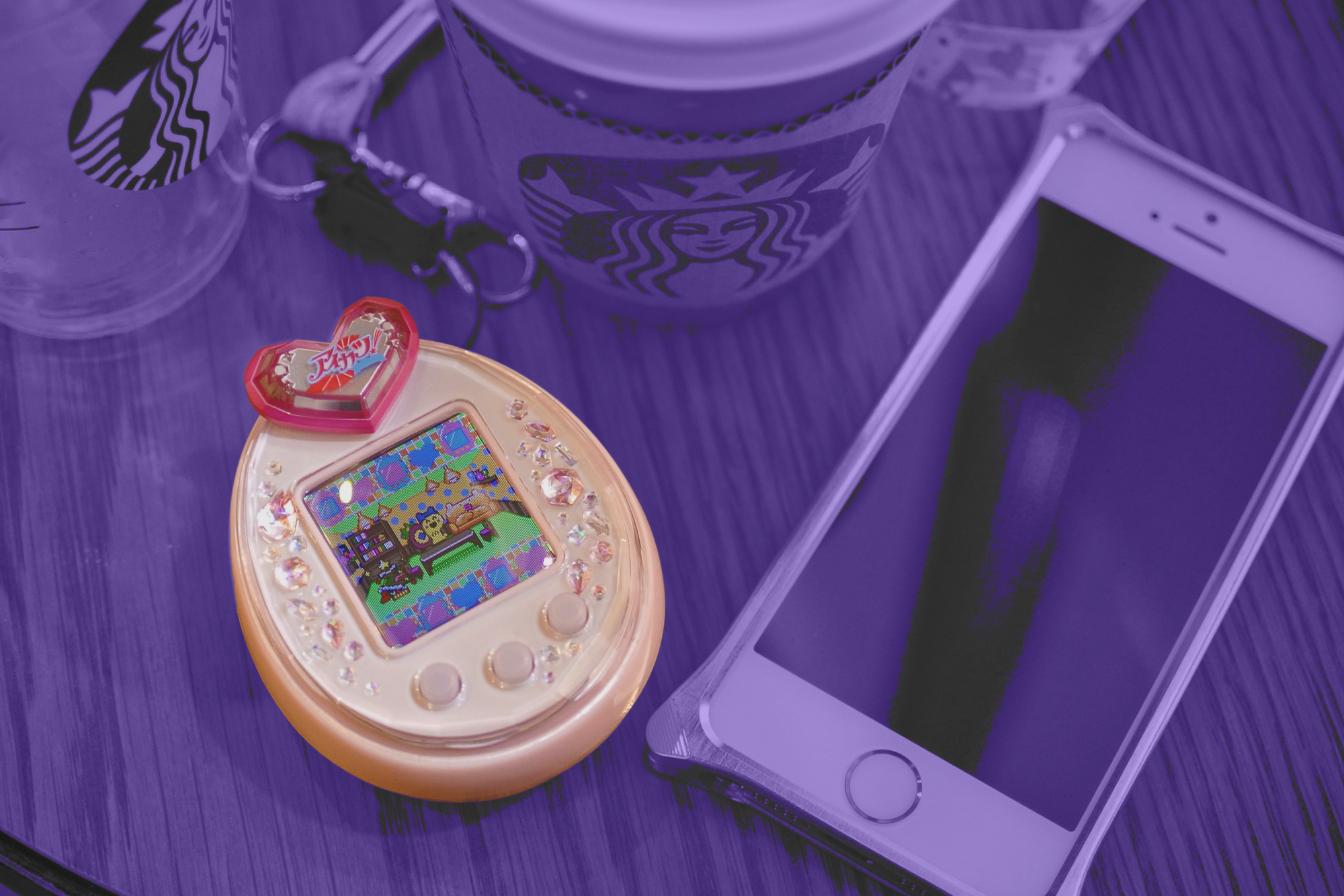 Image of a Tamagotchi next to a mobile phone, treated with a purple filter.