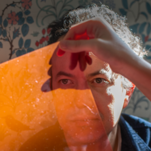 Photograph of Andrew looking through a pane of orange glass, which is being held up by another person's hand.