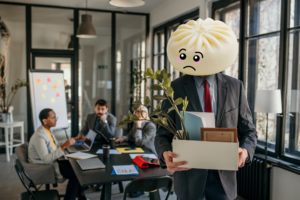 A businessman with the Bun bao for a head carrying his box of items away from an office, clearly just fired. Three people sit at a conference room behind him.
