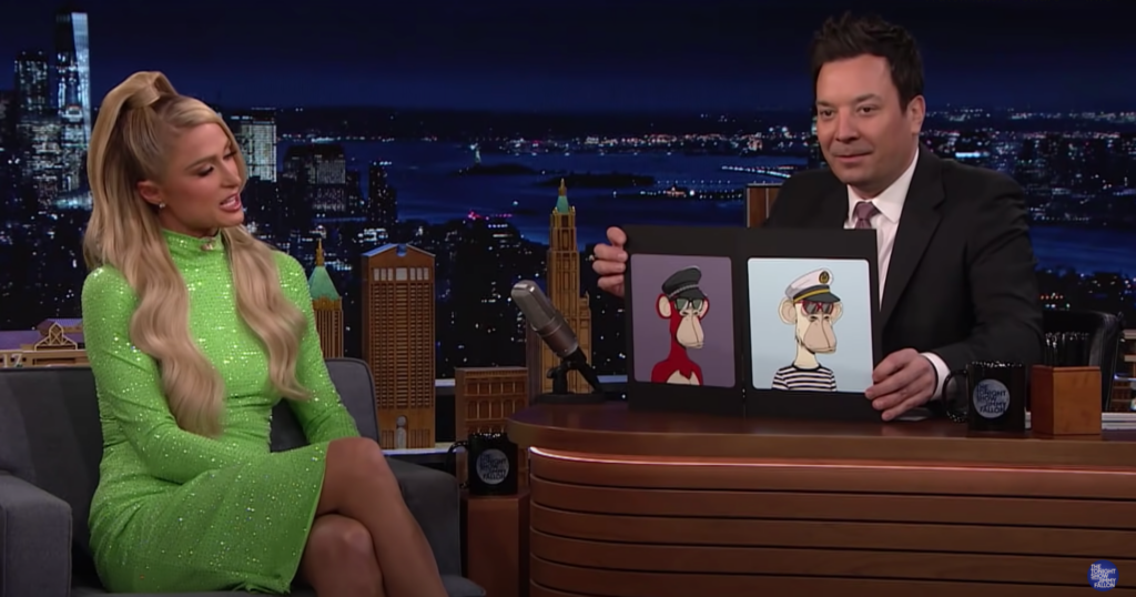 Image from the Tonight Show featuring Paris Hilton in a green dress on the guest couch and Jimmy Fallon behind the host desk. Fallon holds up images of two Bored Apes.